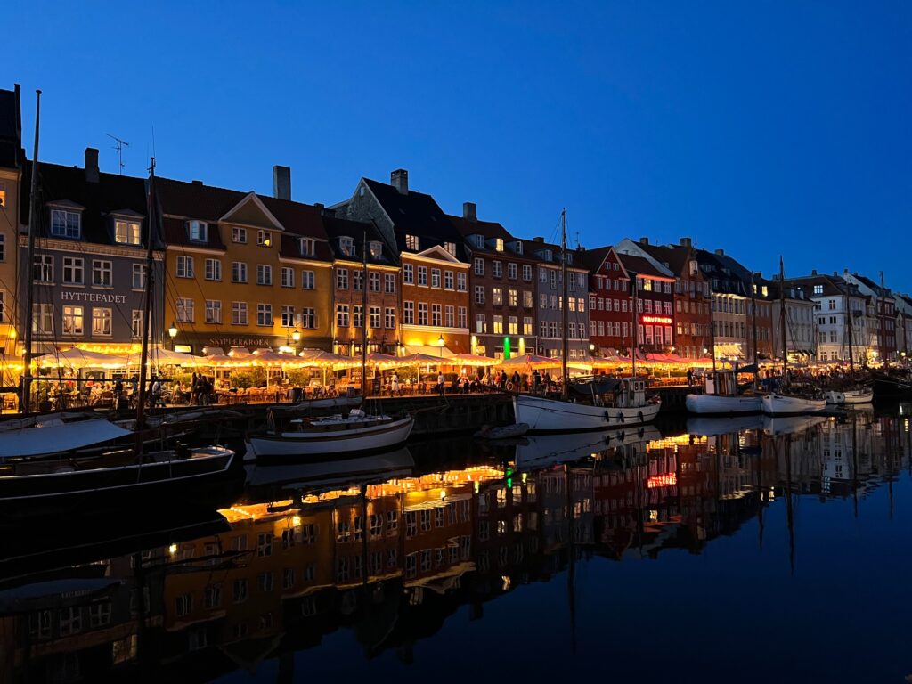 The canal Nyhavn in Copenhagen evening light. Picture taken from the other side of the canal, boats in front and lighted restaurant terraces behind them. In the back colorful old houses.